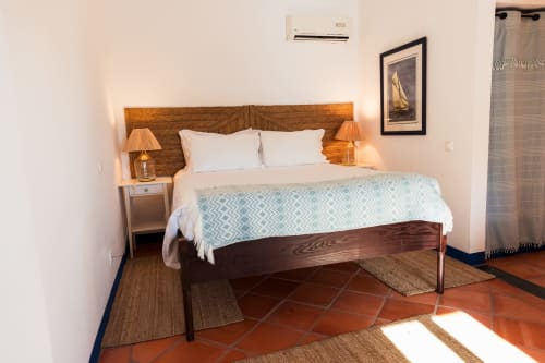 Quarto com cama de madeira, almofadas brancas, manta azul-claro estampada e criados-mudos de madeira com luminárias em Casas de Juromenha