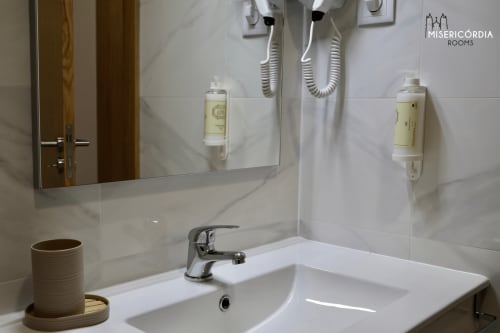 Banheiro moderno da Coleção SM Stays, hotel Guarda, com pia branca, espelho, dois secadores de cabelo e amenidades