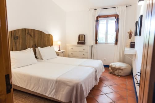 Quarto luminoso na Casas de Juromenha com cabeceira de madeira, roupa de cama branca, móveis creme e azulejos de terracota