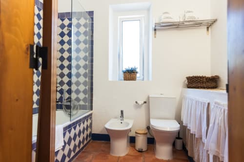 Banheiro da vila Casas de Juromenha em Portugal com chuveiro de azulejos azuis em padrão de diamante, bidê e vaso sanitário brancos, cesto de armazenamento trançado
