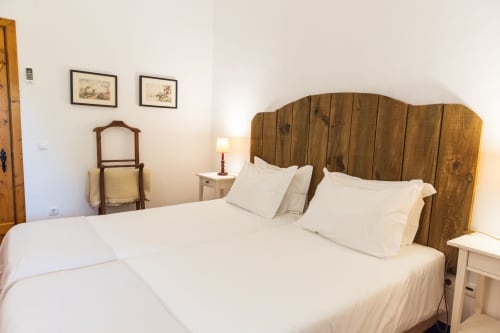 Quarto com cabeceira de madeira, roupas de cama brancas, cadeira rústica e arte emoldurada na villa Casas de Juromenha