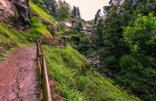 Lush mountainside path with wooden railing, stream, and pink-roofed houses nestled in verdant Portuguese countryside