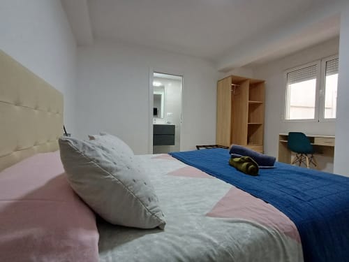 Spacious bedroom at Algemesi Home with blue bedding, white headboard, wooden wardrobe, and ensuite bathroom visible in