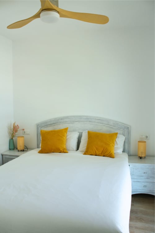 Dormitorio moderno con ventilador de techo de madera, cabecera tapizada gris, ropa de cama blanca y almohadas doradas