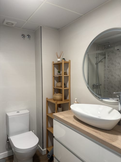Baño moderno con lavamanos blanco tipo recipiente, espejo redondo, estantería de madera, inodoro y cabina de ducha en Love Rent4 Málaga