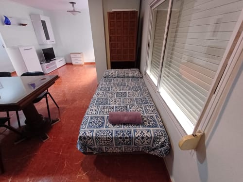 Chambre de l'appartement Algemesi Home avec lit à motifs, carrelage rouge, bureau et portes blanches à volets s'ouvrant sur la terrasse