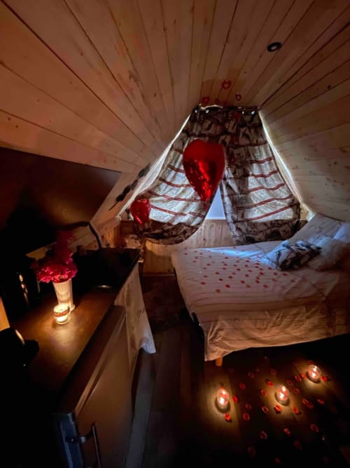 Intérieur romantique de cottage avec lit à baldaquin, guirlandes lumineuses rouges, plafond à poutres apparentes et chandelles aux Les Cabanes