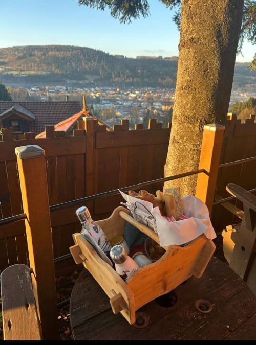Panier de pique-nique en bois garni sur la terrasse avec vue sur la vallée de Granges-Aumontzey et la forêt aux Cabanes des Marchous