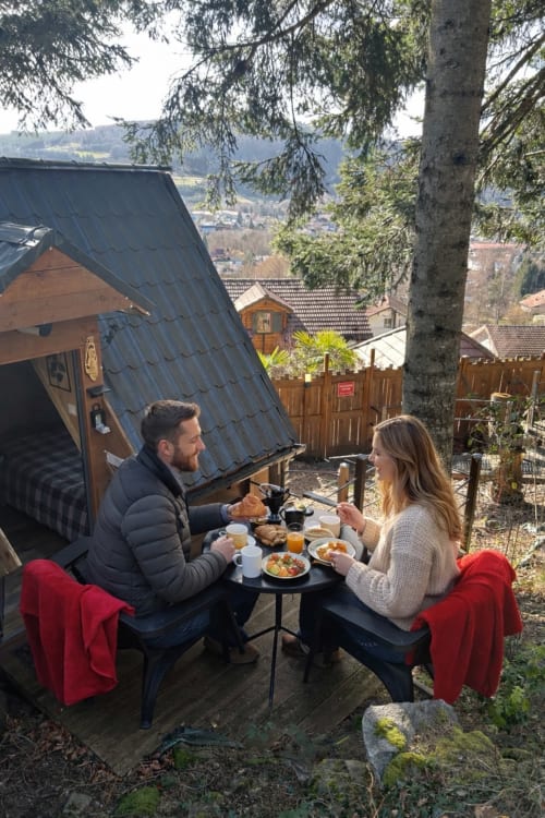 Couple dining outdoors at Cabane Marmotte in Granges-Aumontzey with mountain views and forest backdrop.
