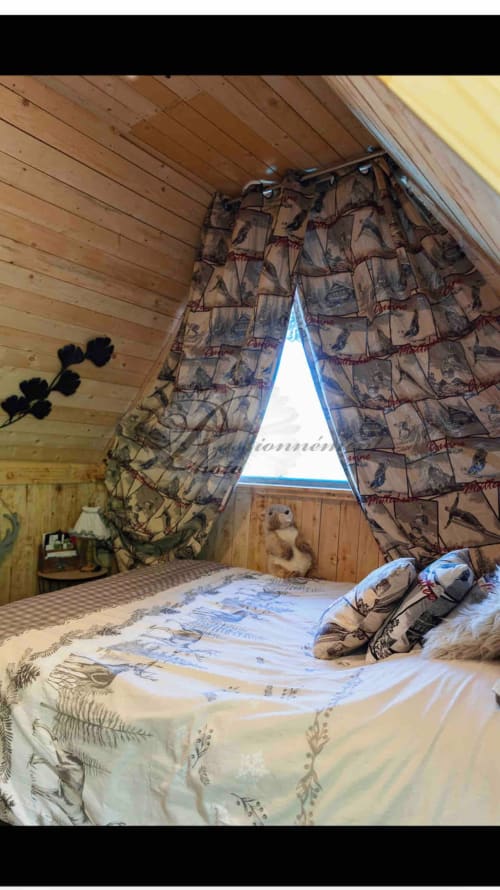 Cozy bedroom in Cabane Marmotte at Les Cabanes des Marchous with triangular window, patterned curtains, and rustic wooden