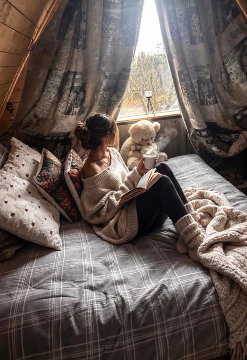 Woman relaxing by window in cozy cottage bedroom with teddy bear, reading and holding warm beverage at Les Cabanes des