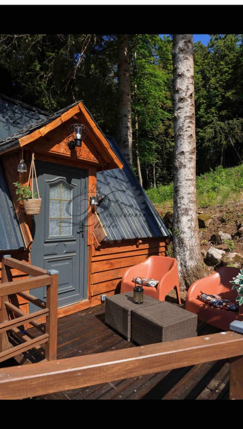 Cozy wooden cottage exterior with orange accents, deck seating, and birch tree at Les Cabanes des Marchous, Granges-Aumontzey