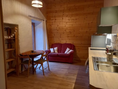 Intérieur douillet de l'Eco Gîte du Mandala avec murs en bois, canapé bordeaux, table à manger et kitchenette avec vue montagne
