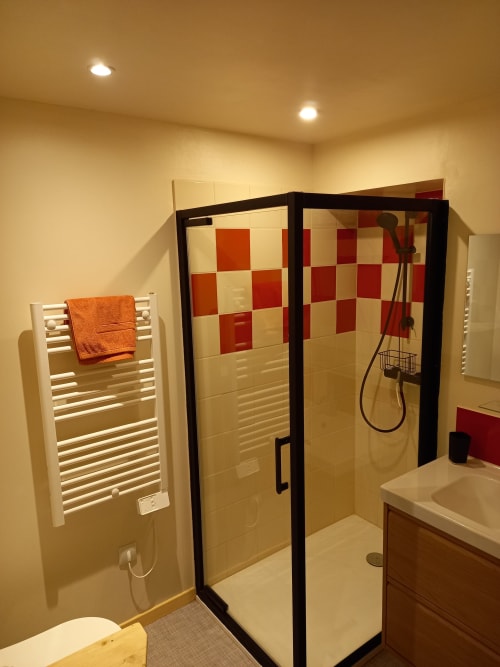 Salle de bain moderne avec douche carrelée rouge et blanc, porte-serviettes blanc avec serviette orange, et radiateur chauffant
