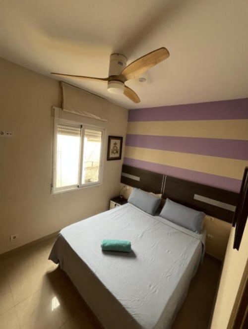 Modern bedroom at Algemesi Home apartment with striped purple and yellow accent wall, ceiling fan, queen bed, and window