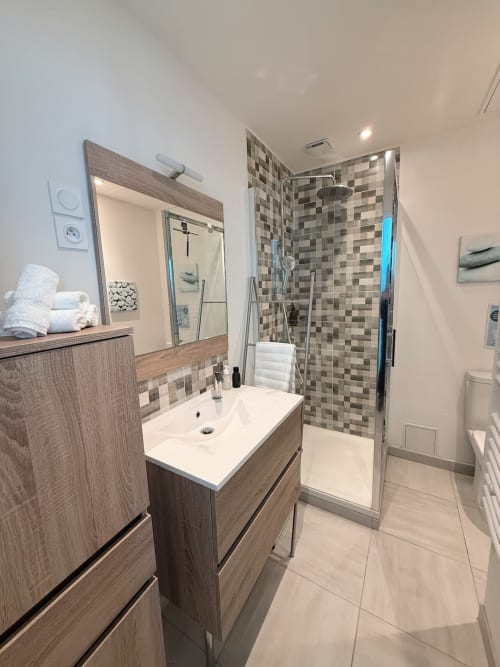 Salle de bain moderne de la villa Maison ARLOÉ à La Rochelle avec meuble vasque en bois, lavabo blanc, douche carrelée en mosaïque et miroir.