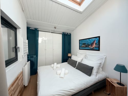 Chambre moderne de la villa Maison ARLOÉ à La Rochelle avec lit blanc, rideaux bleu-canard, verrière et tableau de voilier.