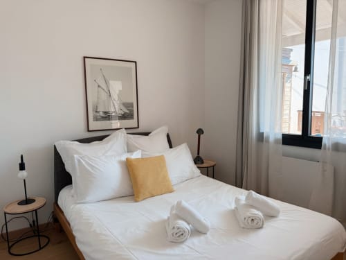 Chambre moderne à la Maison ARLOÉ à La Rochelle avec literie blanche, coussin doré, tableau de voilier et vue sur la ville