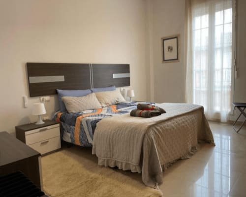 Dormitorio moderno en el apartamento Algemesí Home con cabecero de madera oscura, ropa de cama crema y azul, balcón con vistas a la ciudad