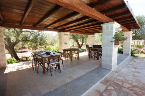 Terrazza coperta con tavoli in pietra, travi in legno e ulivi a Trulli Boccadoro in Puglia