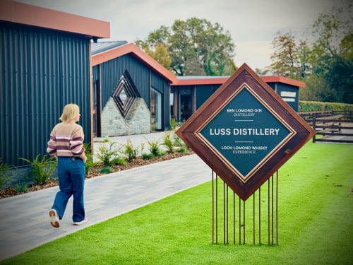 Person approaching Luss Distillery signage at The Corries B&B in Alexandria, Scotland, with modern building and grounds