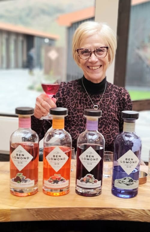Woman with blonde hair and glasses smiling with Ben Lomond gin bottles at The Corries B&B in Luss, Alexandria