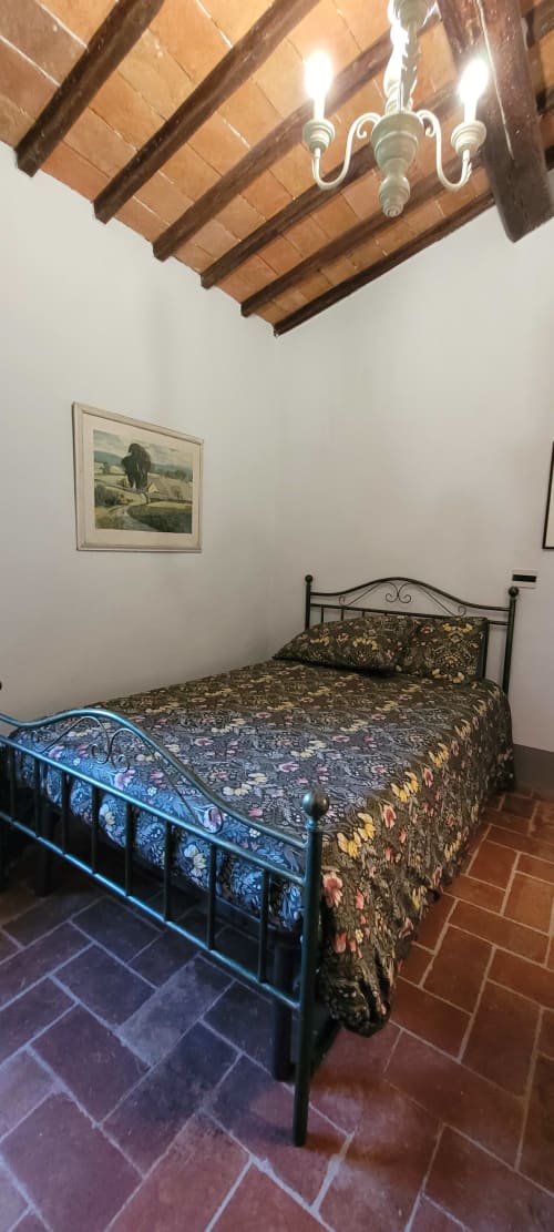 Rustic bedroom with wrought iron bed, exposed wooden beams, terracotta tile floor, and framed countryside art at Rendola