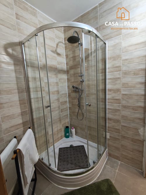 Box de vidro curvado com portas modernas, azulejos estilo madeira bege, chuveiro cascata e tapete preto na Casa Dos Passos