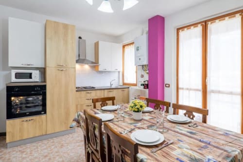 Modern kitchen-dining area with wooden cabinets, dining table set for guests, and pink accent pillar in Como apartment