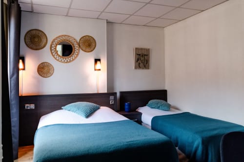 Lits jumeaux avec parure bleue dans une chambre de l'Hôtel les Initiés Rouen, décor mural tissé et éclairage ambiant