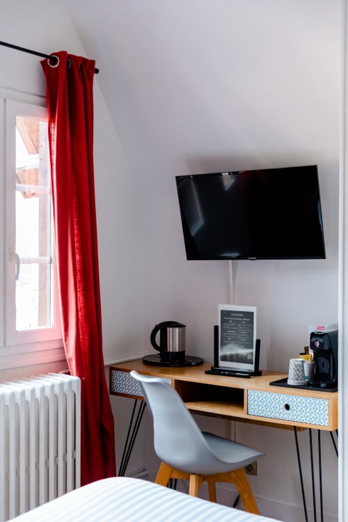 Espace de travail moderne à l'Hôtel les Initiés à Rouen avec bureau, chaise, télévision murale et rideaux rouges près de la fenêtre