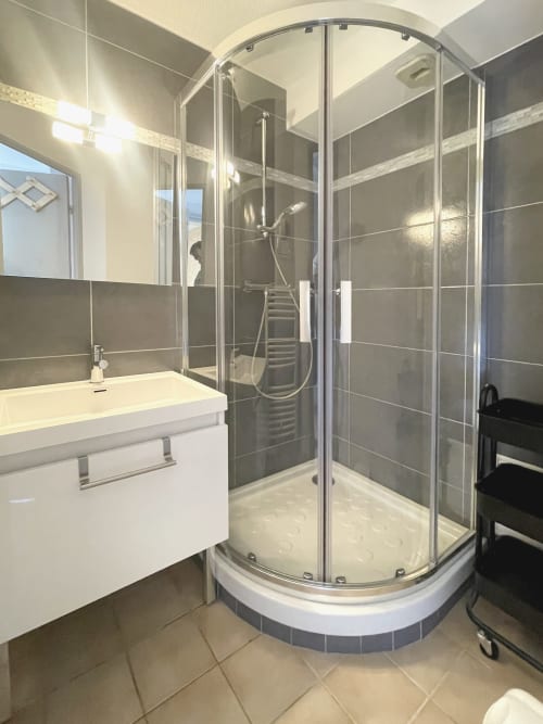 Salle de bain moderne avec douche aux parois courbes en verre, vasque blanche et murs carrelés gris au Vallon des Sources,
