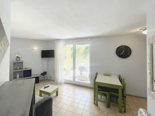 Spacieux séjour lumineux avec mobilier vert, horloge murale, téléviseur et portes vitrées donnant sur la terrasse avec vue montagne au