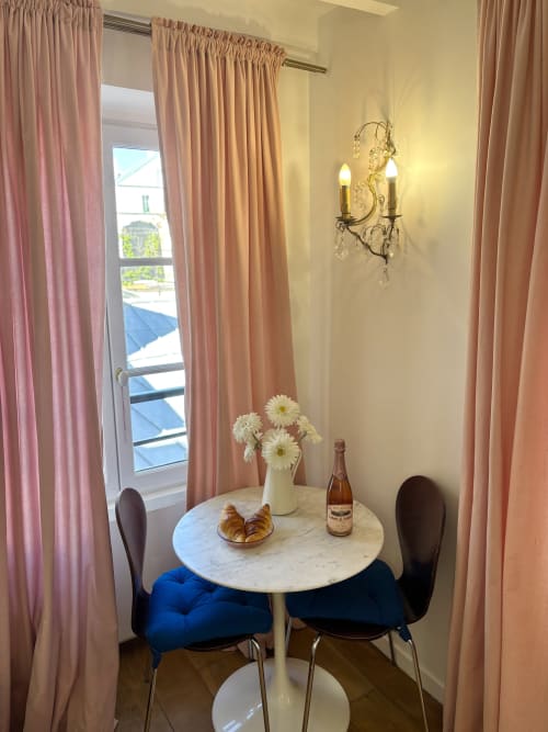 Rincón de comedor elegante en apartamento parisino con mesa de mármol, sillas azules, margaritas blancas, botella de champagne y cortinas rosas