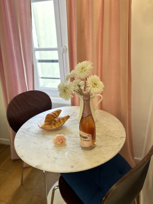 Elegante mesa de comedor de mármol en apartamento parisino con champagne, croissants y flores frescas junto a ventana con cortinas color coral