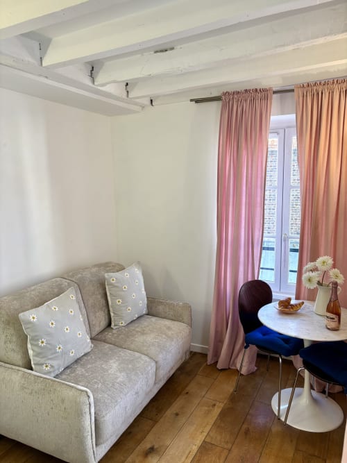 Acogedor salón de apartamento parisino con sofá gris, cojines de margaritas, cortinas rosa, mesa redonda con flores y parquet