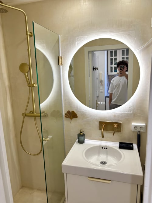 Baño moderno con espejo redondo iluminado, lavabo circular blanco y accesorios de latón en apartamento parisino