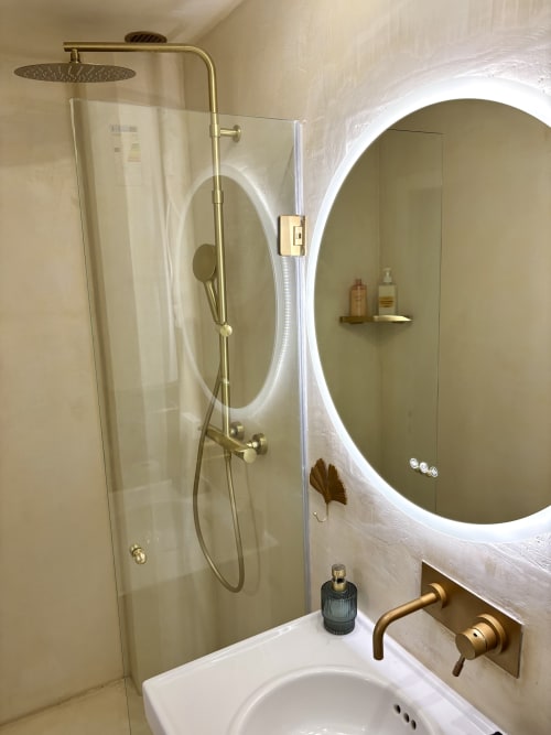 Baño moderno en apartamento parisino con espejo ovalado iluminado, grifería de latón, cabina de ducha de cristal y lavamanos blanco.