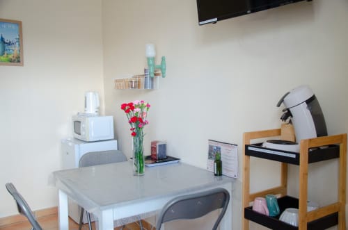 Petite cuisine moderne avec table blanche, chaises grises, œillets roses, cafetière, micro-ondes et étagères en bois aux