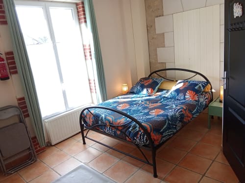 Chambre lumineuse aux Relais-Orbigny avec lit en métal, literie colorée à motifs, carrelage en terre cuite et vue sur le jardin