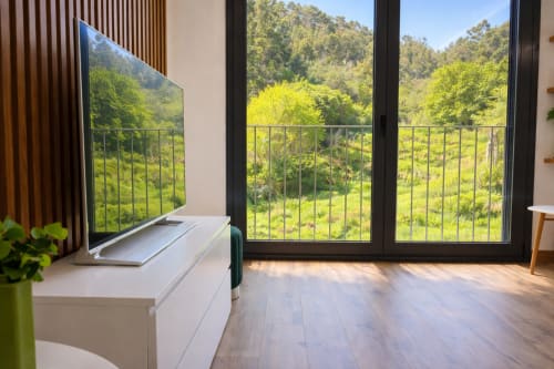 Balcón moderno de apartamento con vistas a montañas en Baiona, España. TV, suelo de madera, amplias ventanas con vistas a vegetación exuberante