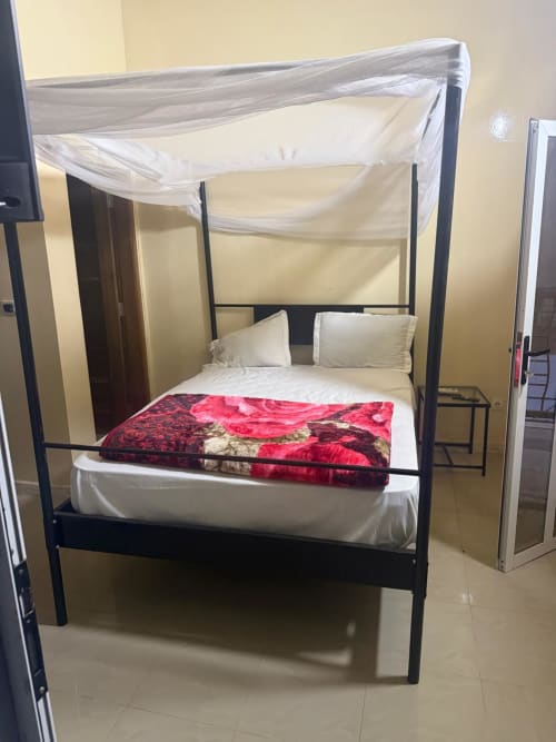 Lit à baldaquin avec voilages blanc crème, couverture rouge motifs, et oreillers crème dans une chambre d'hôtel