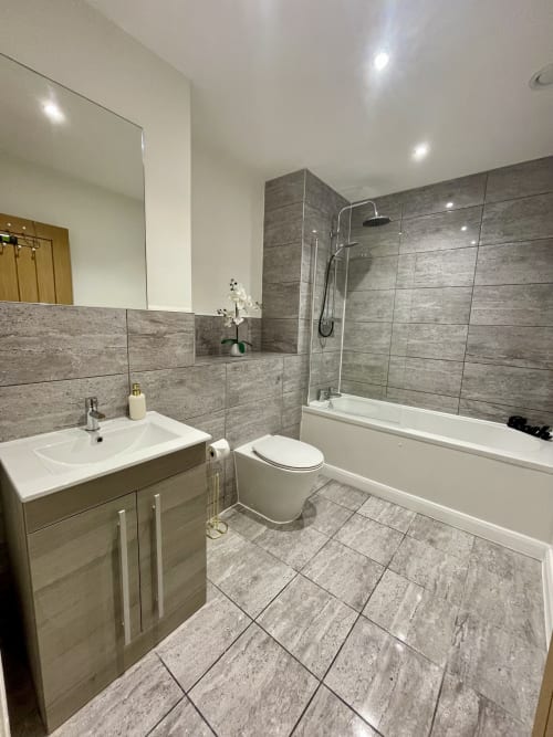 Modern bathroom in Leicester serviced apartment with grey tile walls, white sink vanity, toilet, and shower bathtub combo