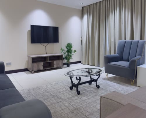 Modern living room at Cozy Apartments Ghana with TV, blue armchair, glass coffee table, and garden views through curtains.