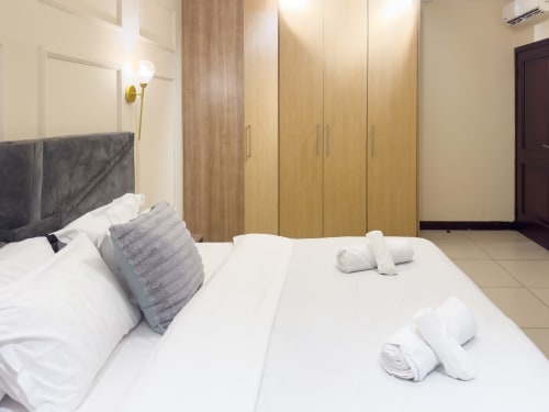 Modern bedroom at Cozy Apartments Ghana with queen bed, grey headboard, wooden wardrobe, white towels, and tile flooring.