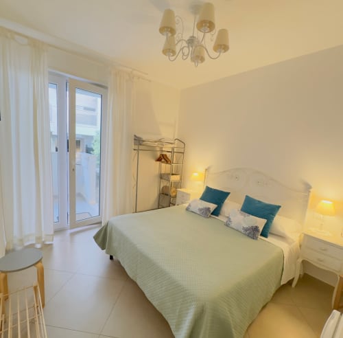 Bright bedroom at B&B Alba in Polignano a Mare with white bed, teal pillows, garden view balcony, and warm lighting.