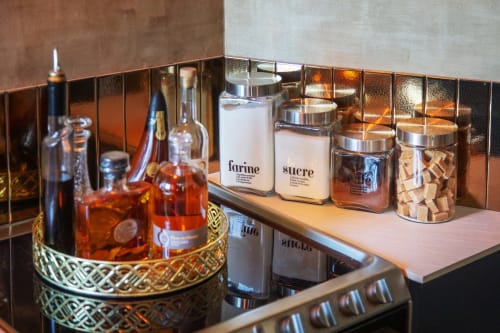 Équipements de cuisine luxe à l'Alombre BnB : bocaux de rangement étiquetés, bouteilles d'alcool, et dosseret décoratif en cuivre