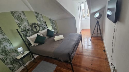 Quarto moderno de hostel em Lisboa com cabeceira de folhas verdes, cama escura, TV, luminária de tripé e pisos de madeira com vista para a cidade