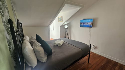 Aconchegante quarto de hostel em Lisboa com cama preta, almofadas brancas, TV exibindo vistas da cidade, piso de madeira e teto inclinado