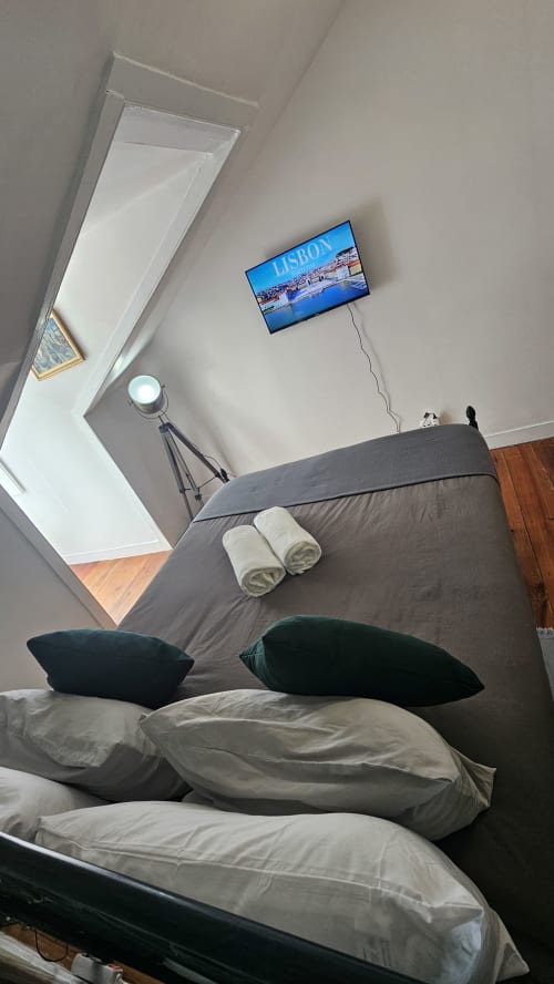Quarto moderno de hostel em Lisboa com roupa de cama cinza, almofadas pretas, TV de tela plana exibindo paisagem urbana de Lisboa, luminária com tripé